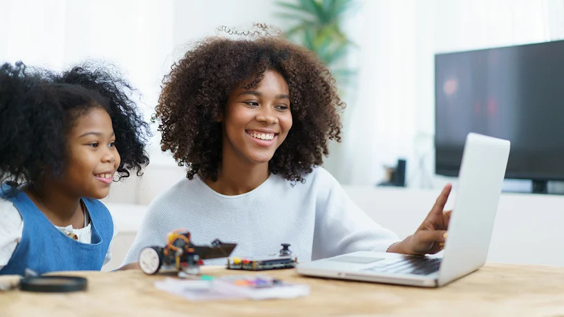Two smiling Black girls, an older sister and a younger one, working together on a laptop for a STEM project. A small toy robot and electronics are visible on the wooden table in front of them