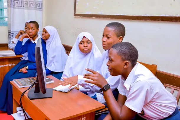 A group of female students in school uniforms, some wearing hijabs, gathered around a computer monitor in a classroom, engaged in collaborative learning.