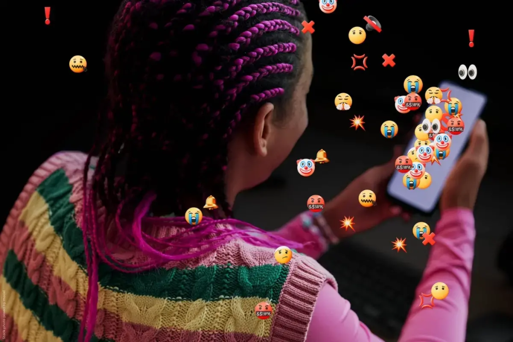 A young woman looks at her smartphone as a swarm of hostile emojis and symbols—including angry faces and exclamation marks—float toward her, illustrating the intensifying digital violence and online abuse facing women and girls.