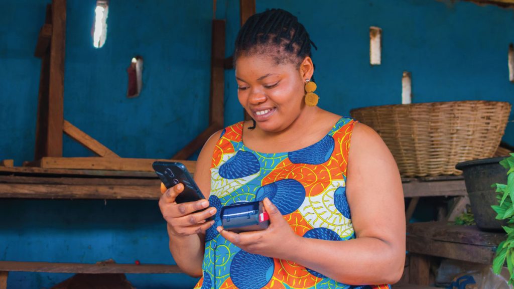 A smiling woman digital entrepreneur uses a smartphone and a mobile point-of-sale (POS) device to manage her business, symbolizing the journey of breaking down barriers for women in the digital economy.
